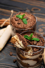 Classic tiramisu dessert in a glass cup, pieces of chocolate and savoiardi cookies on wooden background