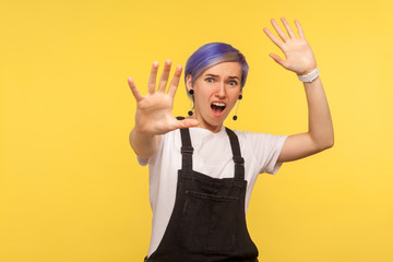 Portrait of scared hipster woman with violet short hair in denim overalls showing stop gesture and looking surprised frightened, shouting from sudden fear. isolated on yellow background, studio shot