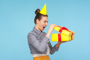 Extremely surprised emotional woman with party cone on head opening gift and looking inside box,...