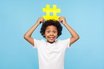 Funny excited surprised little boy with curly hair in T-shirt holding big yellow hashtag symbol over his head and looking with amazement, popular blogging, child content. studio shot n blue background