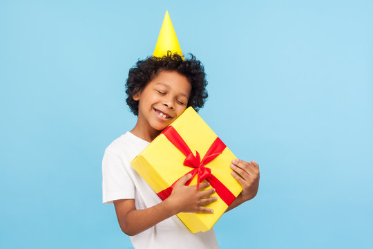 Amusing Happy Pleased Little Boy With Party Cone On Head Embracing Gift Box And Closing Eyes In Pleasure, Child Enjoying Long-awaited Birthday Present, Dreams Come True. Studio Shot Blue Background