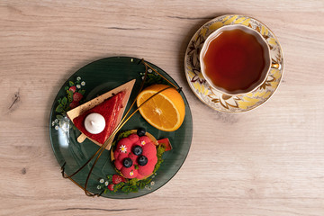 Two appetizing delicious red cakes and half of juicy orange on a glass plate with cup of tea nearby, top view