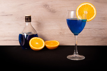 Glass with blue beverage and slice of juicy orange on the side, spilled bottle and orange halves behind
