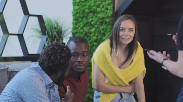 nice girl in yellow sweater on shoulders sits with African guy friends on couch and makes order to waiter close up slow motion and shows dissatisfaction with bill.