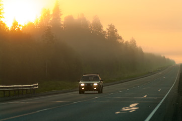 Passenger car driving through the fog at morning time, sunshine with orange sky, empty road going to distance