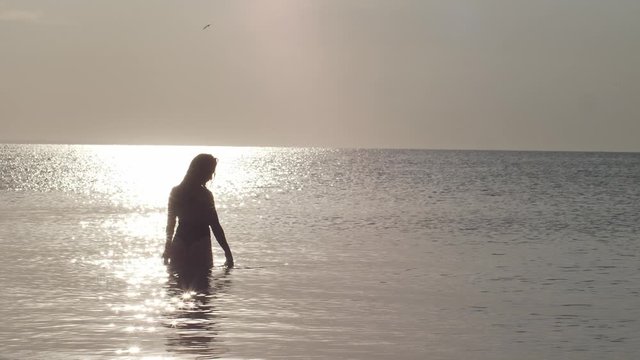 A beautiful woman in a swimsuit stands in the middle of a calm sea at dawn. The sun's rays and glare play on the water shining through the female image.