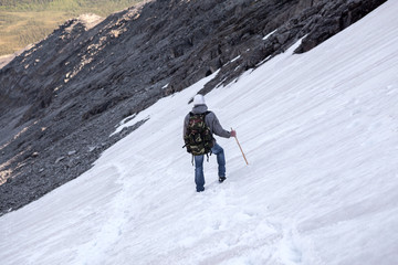 Hiker climbs down a mountain, walks on snowy steep slope with stick, descent into a valley