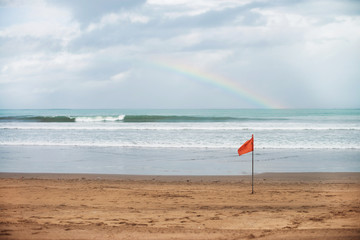 Beautiful sea landscape: summer beach with rainbow in cloudy sky,  storm waves and red beach flag. After the rain. Storm and tourism concept. 