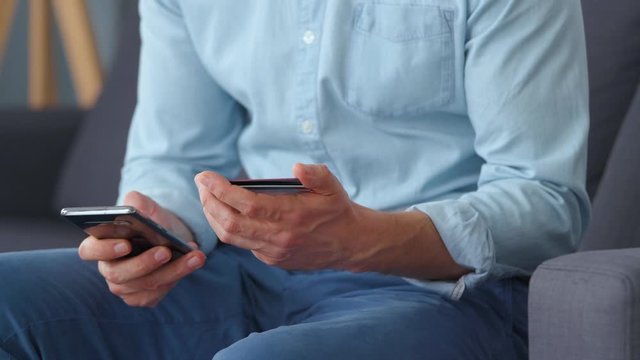 Formally dressed man sits on a sofa and enters a credit card number into a smartphone to pay online. Online shopping, lifestyle technology. Close-up