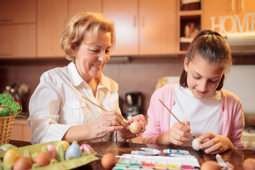 Smiling senior woman decorating and painting Easter eggs together with her teenage granddaughter in...