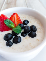 Oatmeal with sliced strawberries and blueberries in a white bowl decorated with mint leaf. Selective focus