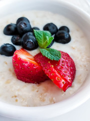 Oatmeal with sliced strawberries and blueberries in a white bowl decorated with mint leaf. Selective focus