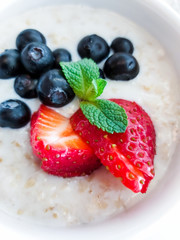 Oatmeal with sliced strawberries and blueberries in a white bowl decorated with mint leaf. Selective focus