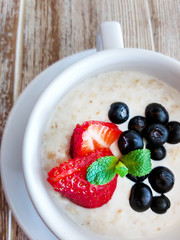 Oatmeal with sliced strawberries and blueberries in a white bowl decorated with mint leaf. Top view