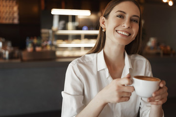 Fototapeta na wymiar Close-up portrait happy alluring lady in white blouse, smiling cheerfully as sitting in cafe, businesswoman drinking coffee, relaxed rejoicing woman winning deal in court, have break