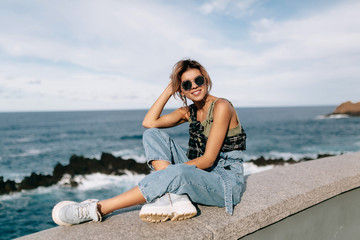 Full-lenght outside portrait of stylish european woman sitting by the ocean and blue sky