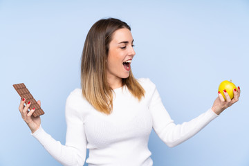 Young blonde woman over isolated blue background taking a chocolate tablet in one hand and an apple...
