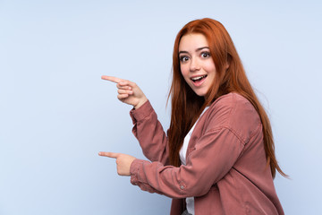 Redhead teenager girl over isolated blue background surprised and pointing side