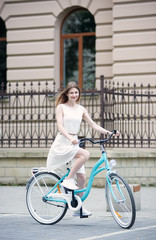 Fototapeta na wymiar Young woman in light white dress is riding a blue bike on the street in the city smiling and looking at the camera, full length, beautiful building with fence on background
