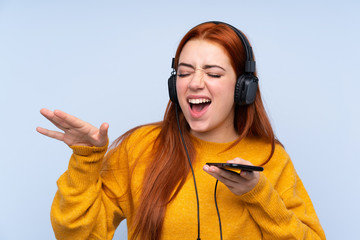 Redhead teenager girl over isolated blue background listening music with a mobile and singing