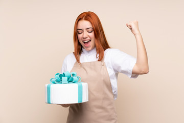Redhead teenager girl with a big cake over isolated background celebrating a victory