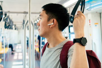 Close up of young man listening to music with wireless earpods while commuting by train. Asian guy...