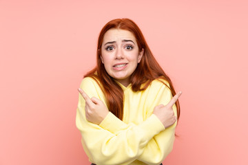 Redhead teenager girl over isolated pink background pointing to the laterals having doubts