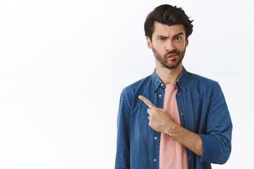 Skeptical, uncertain and suspicious handsome 25s man with beard, frowning smirk unsure, pointing left and staring doubtful camera, cant decide, standing indecisive over white background