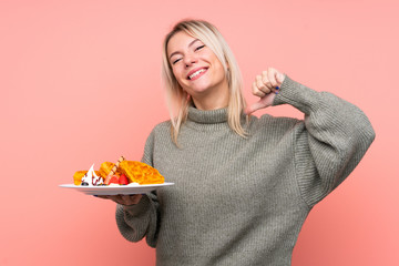 Young blonde woman holding waffles over isolated pink background proud and self-satisfied