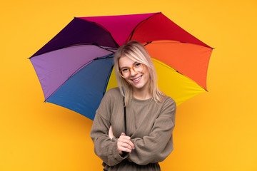 Young blonde woman holding an umbrella over isolated yellow wall laughing