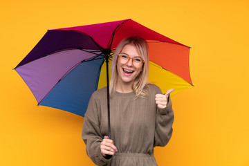 Young blonde woman holding an umbrella over isolated yellow wall giving a thumbs up gesture