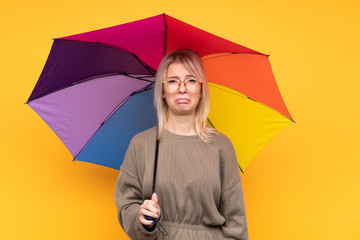 Young blonde woman holding an umbrella over isolated yellow wall sad