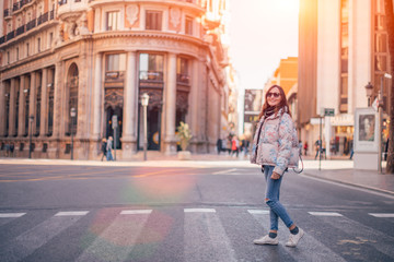 Fototapeta premium Smiling girl walking down the street, crossing the road at a pedestrian crossing. Sunny spring day, image with copy space