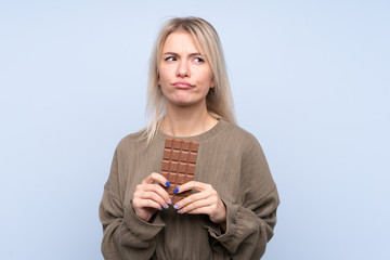 Young blonde woman over isolated blue background taking a chocolate tablet and having doubts