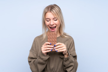 Young blonde woman over isolated blue background eating a chocolate tablet