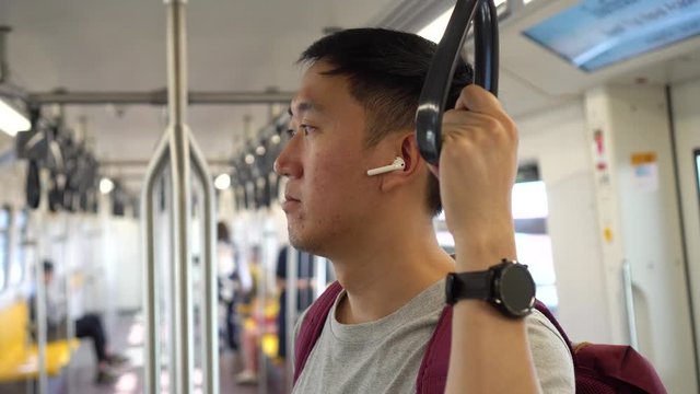 Close up of young man listening to music with wireless earpods while commuting by train. Asian guy enjoying music on the go
