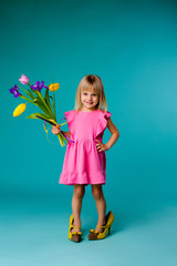 little blonde girl in big yellow shoes and a pink dress is holding spring flowers, baby girl in a pink dress and mother's shoes smiling on a blue background, a little fashionista. space for text