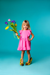 little blonde girl in big yellow shoes and a pink dress is holding spring flowers, baby girl in a pink dress and mother's shoes smiling on a blue background, a little fashionista. space for text