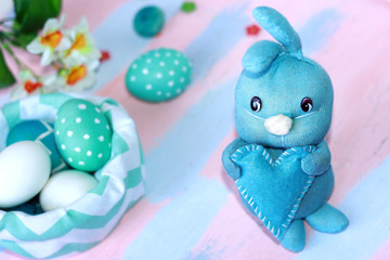 Easter light blue rabbit stands with a blue heart in its paws near the basket with blue-white eggs. Handmade. Greeting card for happy easter.