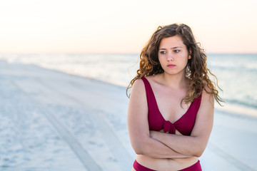 Young woman cold shivering unhappy sad crossed arms in red bikini swimsuit standing in beach sunrise or sunset in Florida panhandle with ocean and sand