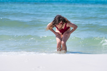 Young woman in swimsuit bikini bathing suit bending down washing legs with water waves splashing on shore in Santa Rosa Beach, Florida