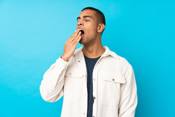 Young African American man over isolated blue background yawning and covering wide open mouth with hand