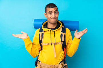 Young mountaineer man with a big backpack over isolated blue background having doubts with confuse...