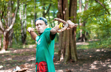 A young man in a green T-shirt and bandana shoots from a large wooden slingshot.