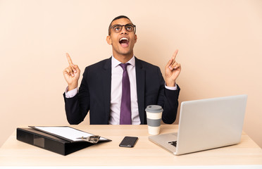 Young business man in his office with a laptop and other documents surprised and pointing up