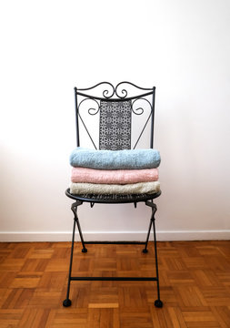 Folded Light Blue, Pink And Grey Bath Towels On The Forged Iron Chair With White Walls And Brown Parquet Floor