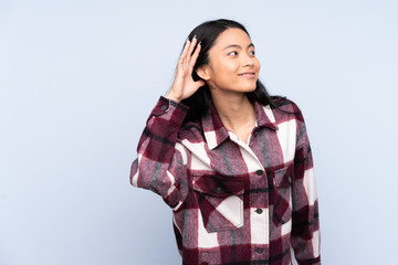 Teenager Chinese woman isolated on blue background listening to something by putting hand on the ear