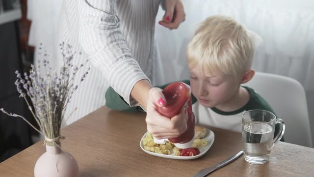 Mom pours ketchup on her son's plate with pasta and meatballs at lunch.