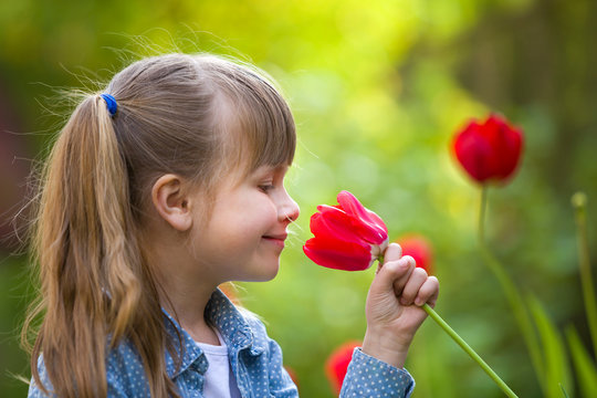 Profile of cute pretty smiling child girl with gray eyes and long hair smelling bright red tulip flower on blurred sunny summer green bokeh background.