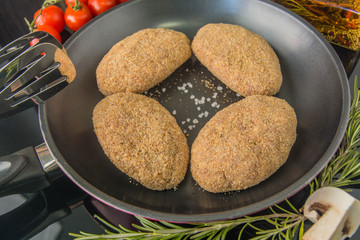 Raw cutlet in breadcrumbs. Frying pan, induction cooker, spices, olive oil, salt, tomato, rosemary, mushrooms, cream plan, mirror surface.